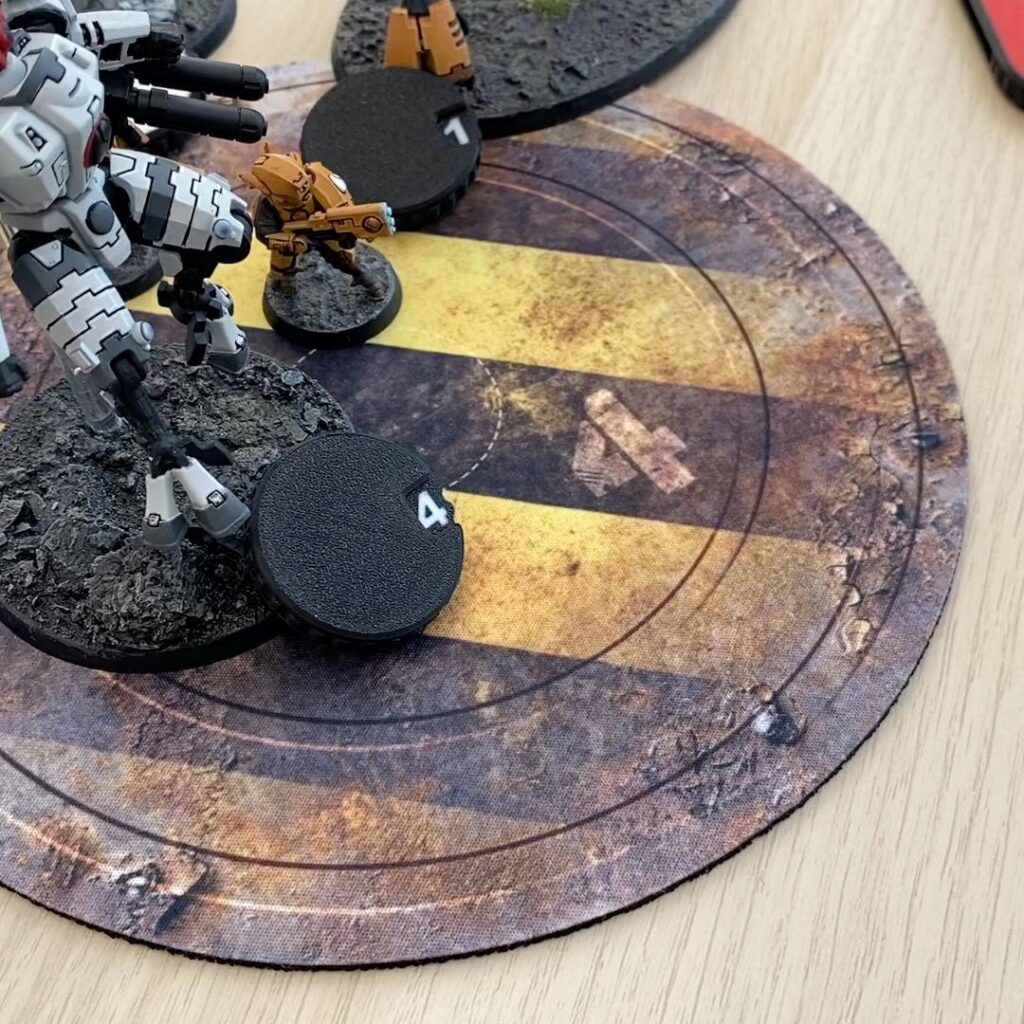 A white and grey mechanical robot miniature stands on a textured base next to a black circular token marked with the number 4.