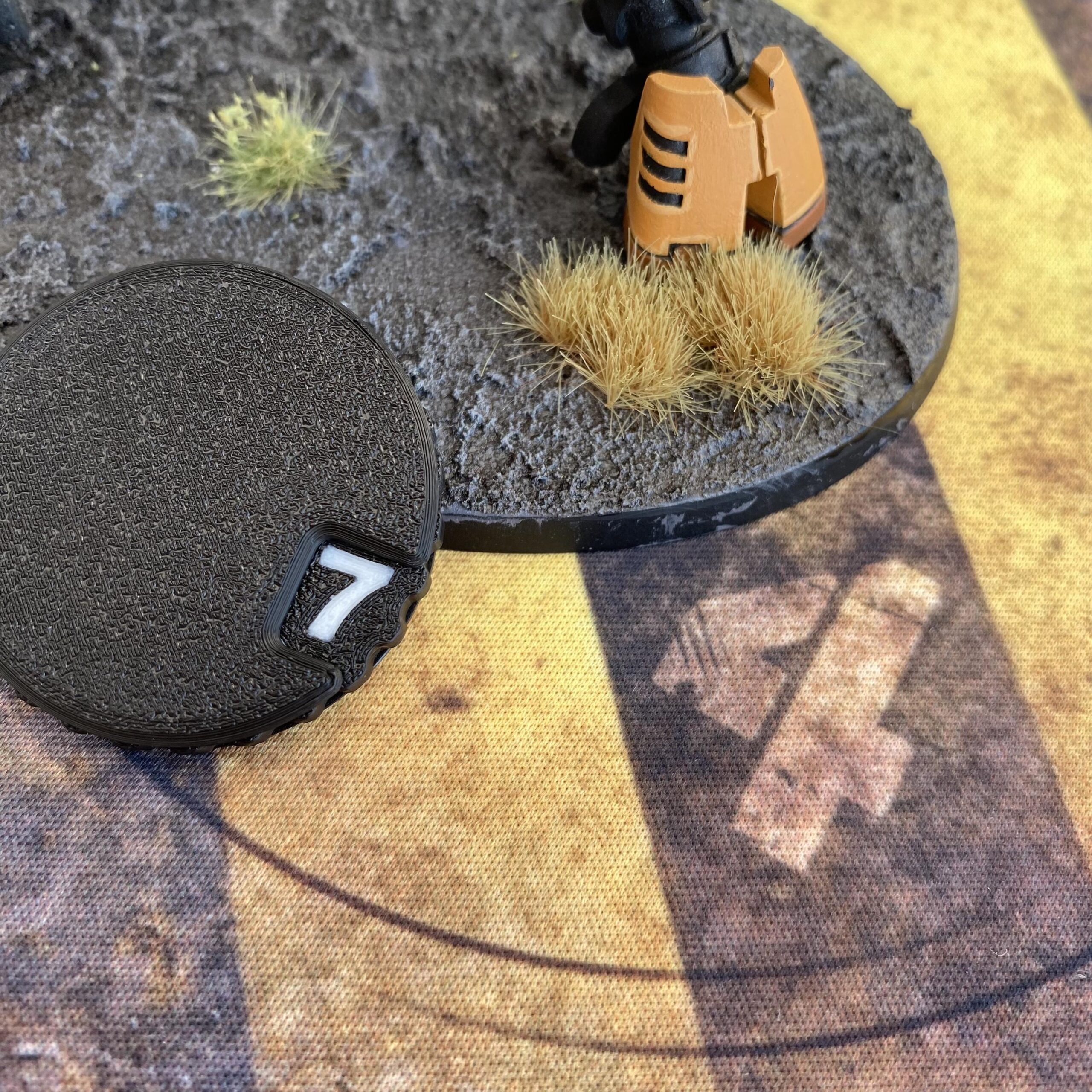 A black circular token with a white number 7 printed on its edge, positioned next to a textured miniature base featuring dark grey terrain, tan tufts of grass, and a partial tan and black mechanical component on the surface.