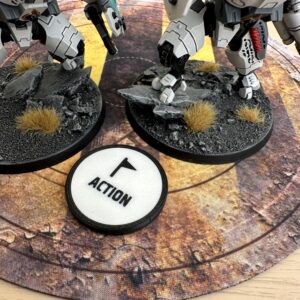 Two white and grey robotic miniature figures stand on circular bases with textured grey rock and brown grass details.