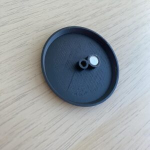 A black, circular object with a raised edge and a small, cylindrical protrusion.